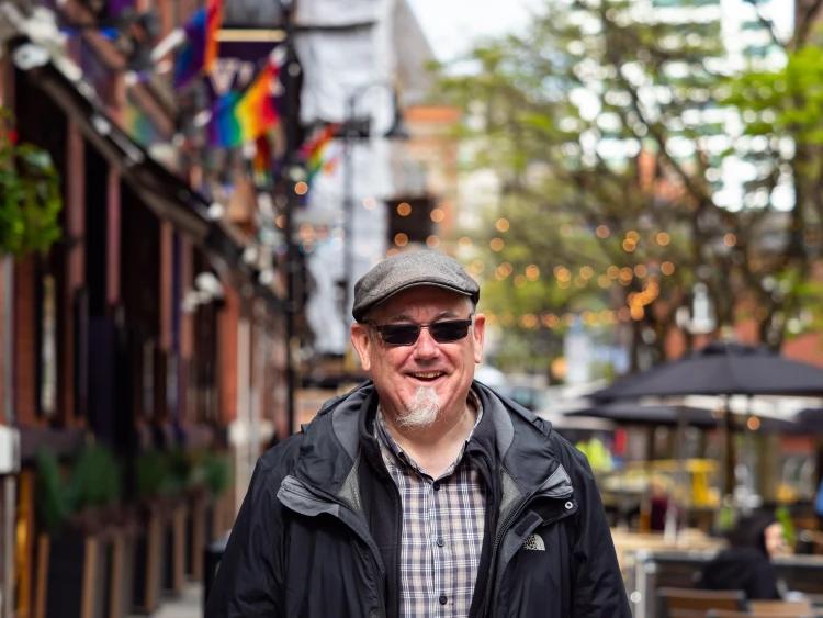 Man in street wearing cap and sunglasses with rainbow flags behind him
